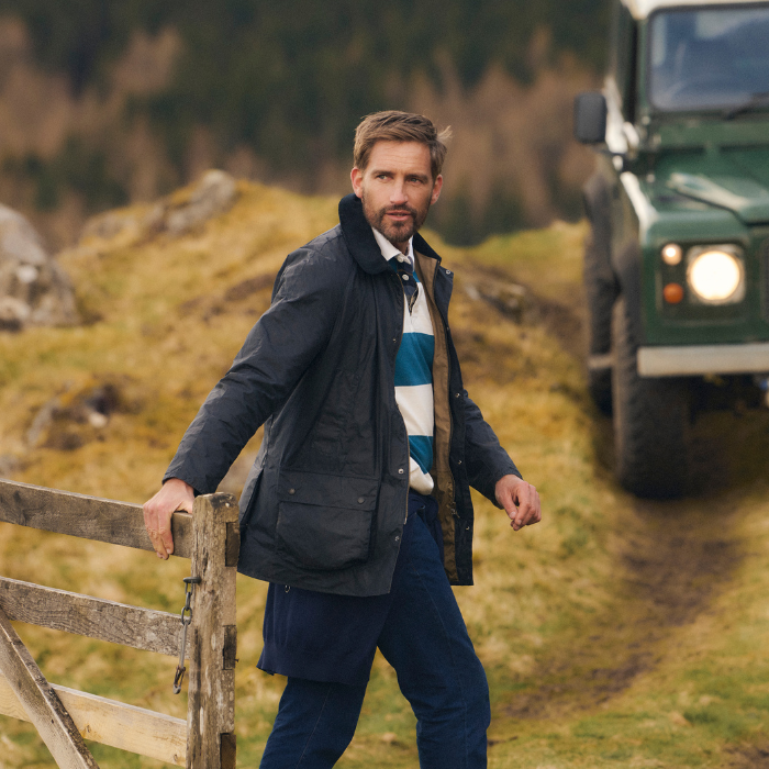 Man closing a gate wearing the Barbour Ashby Lightweight Wax Jacket with a rugged country landscape and green Land Rover Defender in the background