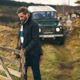 Man manoeuvres a gate wearing the Barbour Ashby Lightweight Wax Jacket with a country landscape and green Land Rover Defender in the background