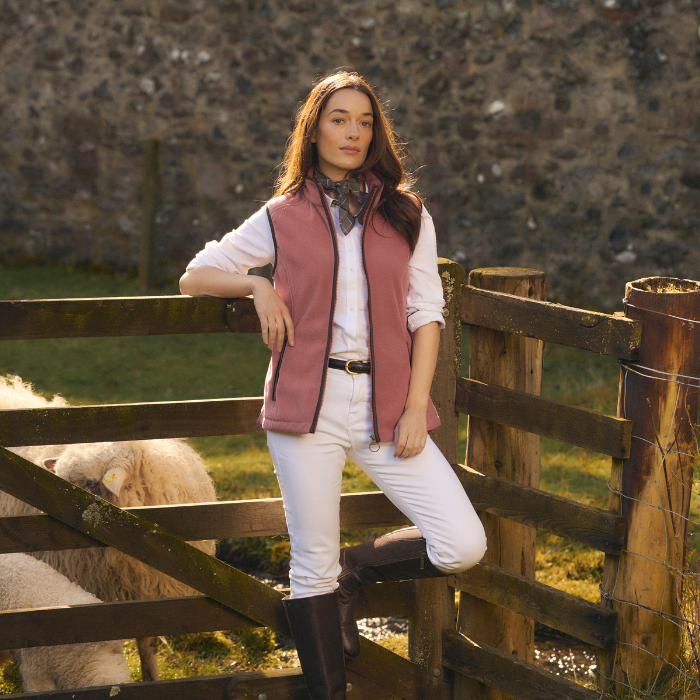 Female leans casually against a sheep enclosure whilst wearing crisp white jeans and shirt, layered over with the Barbour Ladies Country Colton Fleece Gilet in Deep Pruce