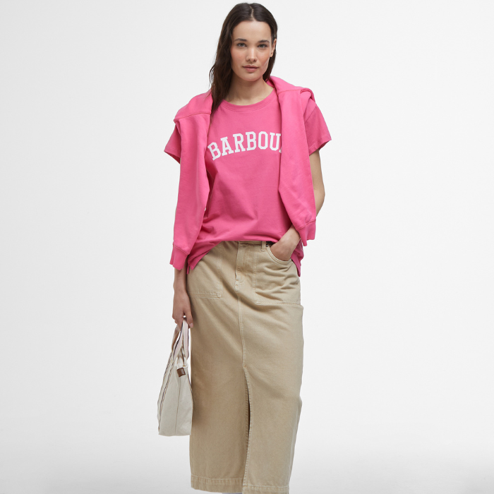 Female styles the Barbour Ladies Northumberland Classic T-shirt in Rose Pink with a long beige skirt and vibrant pink jumper draped across her shoulders