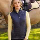 Female in the forefront wearing the LeMieux Ladies Juliette Gilet in Dusk Blue with a horse in the background