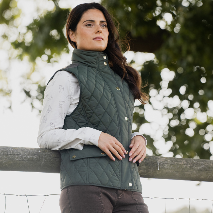 Woman leans against a fence wearing the Toggi Ladies Storford Quilted Gilet in Dark Khaki