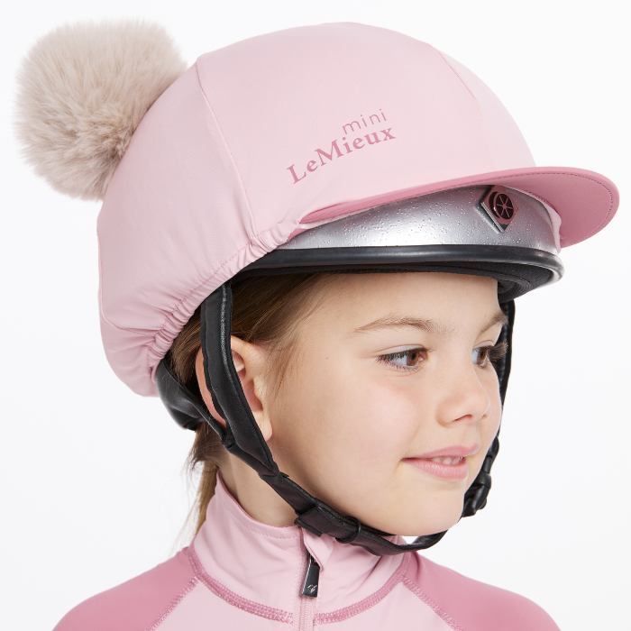 Little girl wears her riding helmet with the LeMieux Mini Double Pom Hat Silk in Blossom